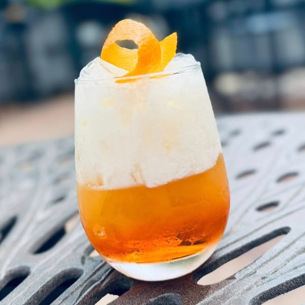 A Label Kitchen + Bar cocktail with orange liquid, white foam, and an orange peel garnish.