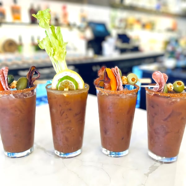 Four Bloody Mary cocktails with various garnishes, including celery, lemon, and charcuterie, on a bar.