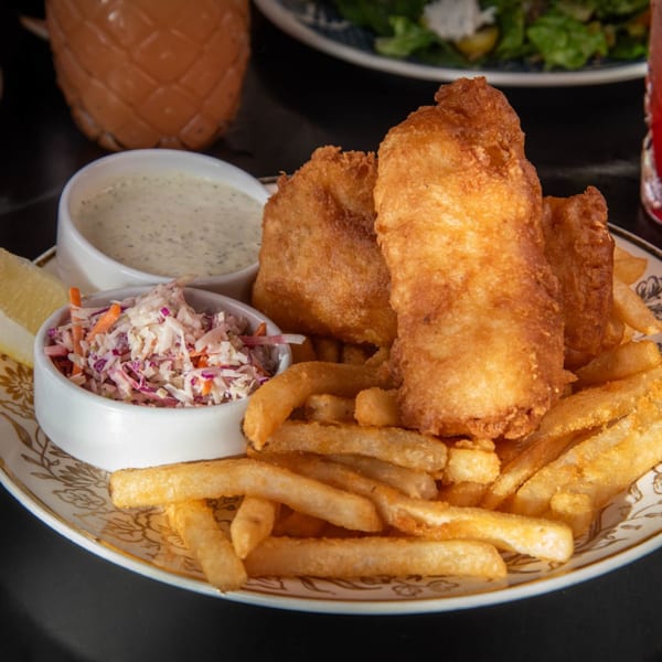 Golden-brown fish and chips with coleslaw, tartar sauce, and a lemon wedge on a decorative plate.