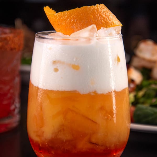 A Label Kitchen + Bar cocktail with orange liquid, white foam, ice, and an orange peel garnish.