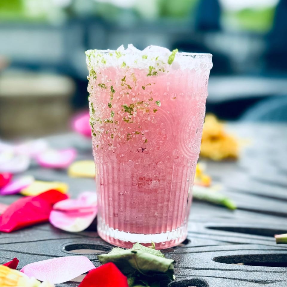 Pink cocktail with a lime-salt rim and ice, surrounded by colorful rose petals on a dark table.