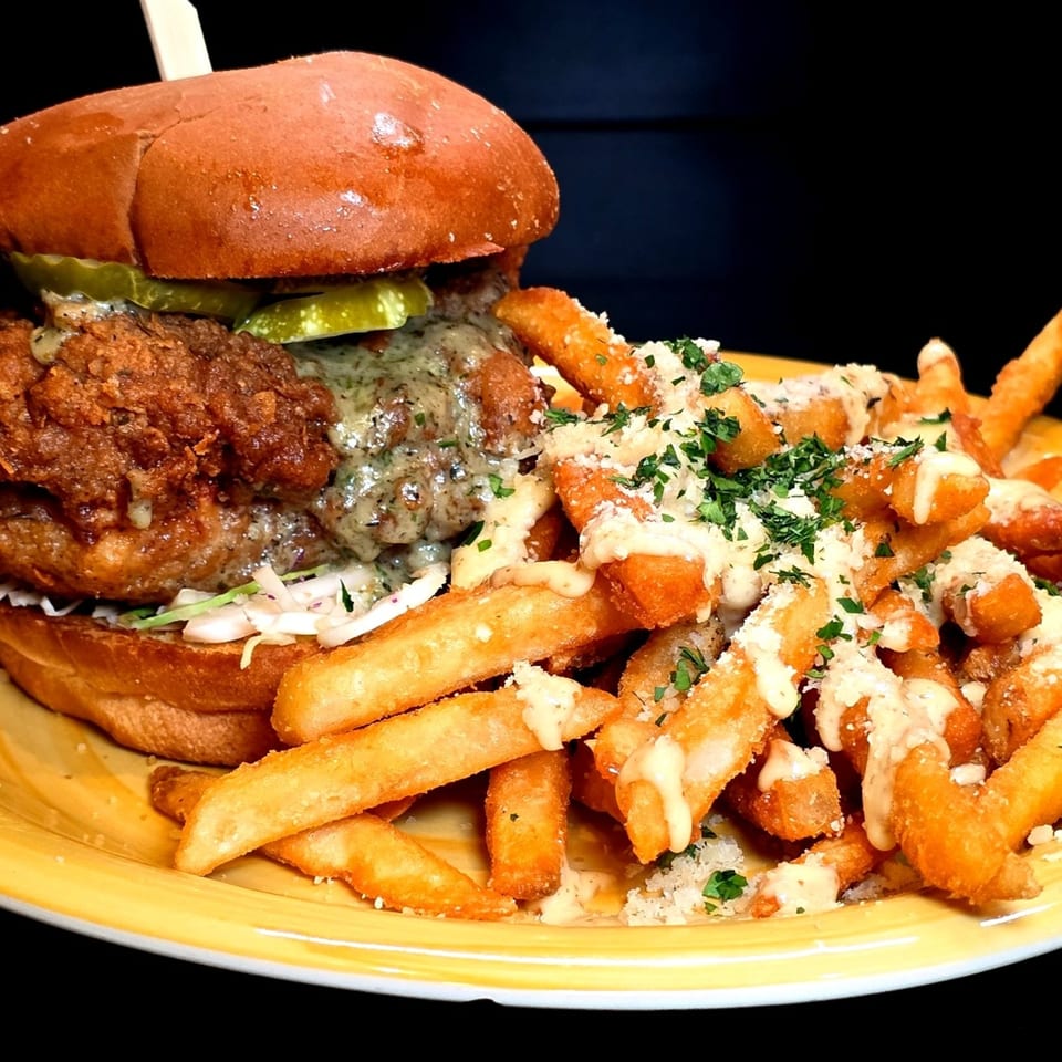 A fried chicken sandwich with pickles and sauce, served with parmesan fries on a yellow plate.