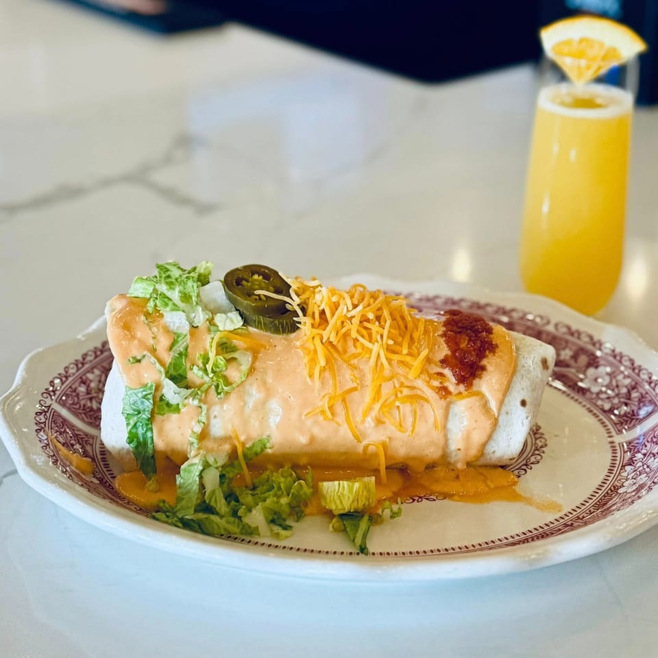 A burrito topped with sauce, cheese, lettuce, and jalapeños, with a mimosa in the background.