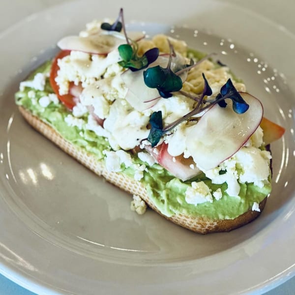 Avocado toast with feta, sliced radishes, tomatoes, and microgreens on a white plate.