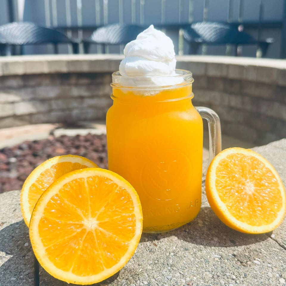 Orange drink with whipped cream in a mason jar, surrounded by orange slices on a stone surface.