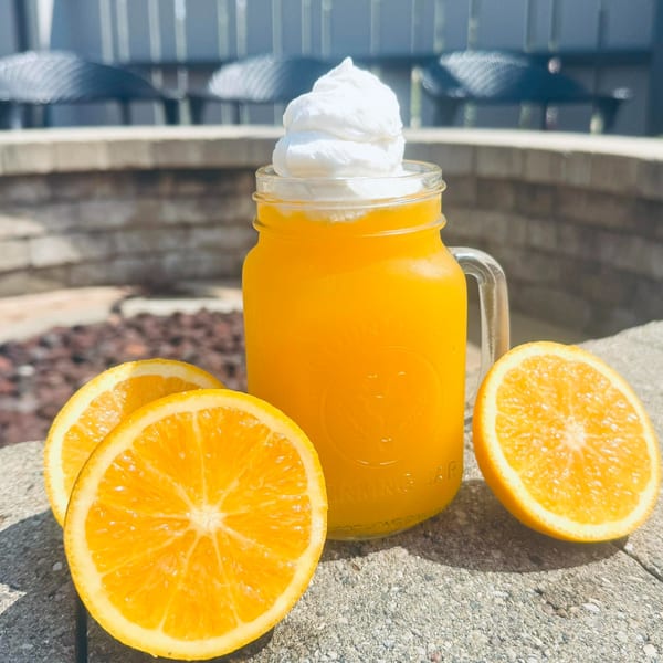 Orange drink with whipped cream in a mason jar, with orange slices on a stone surface.