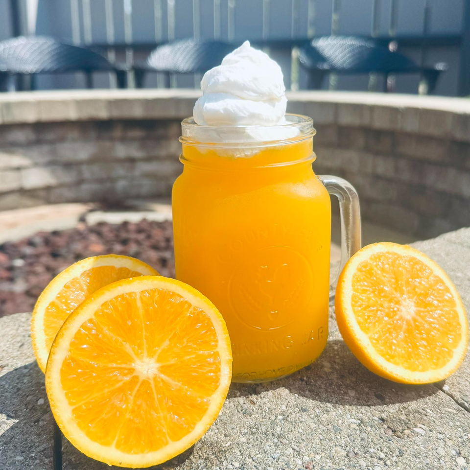 Orange drink with whipped cream in a mason jar, with orange slices on a stone surface.