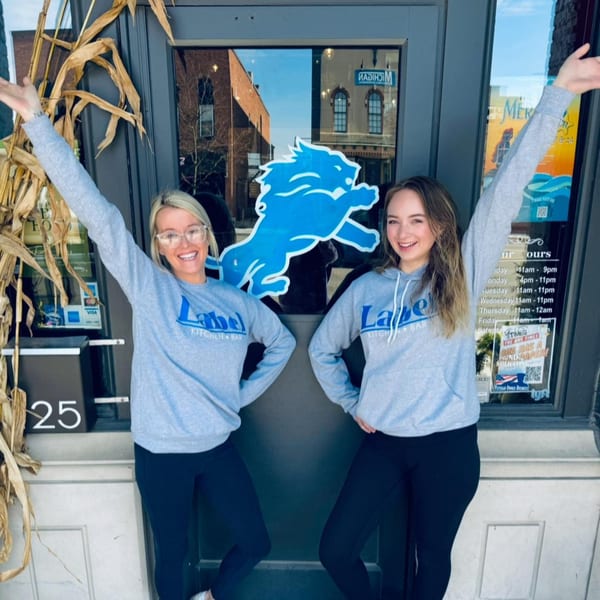 Two women in Label Kitchen + Bar hoodies stand smiling with arms raised in front of the restaurant entrance.