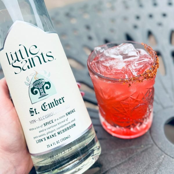 A hand holds a bottle of Little Saints St. Ember non-alcoholic spirit next to a red cocktail.