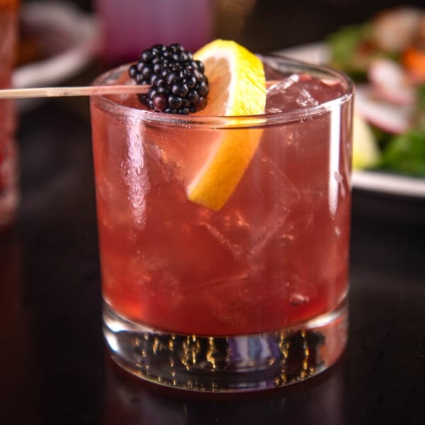 A red cocktail in a rocks glass, garnished with a blackberry skewer and lemon wedge.