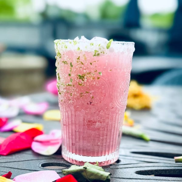 Pink cocktail with a salt and herb rim, surrounded by colorful flower petals on a dark patio table.