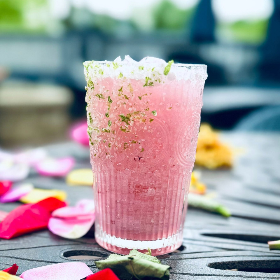 Pink cocktail with a salt and herb rim, surrounded by colorful flower petals on a dark patio table.