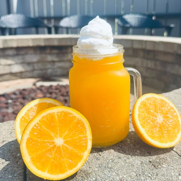 Orange drink with whipped cream in a mason jar, surrounded by orange slices on a stone surface.
