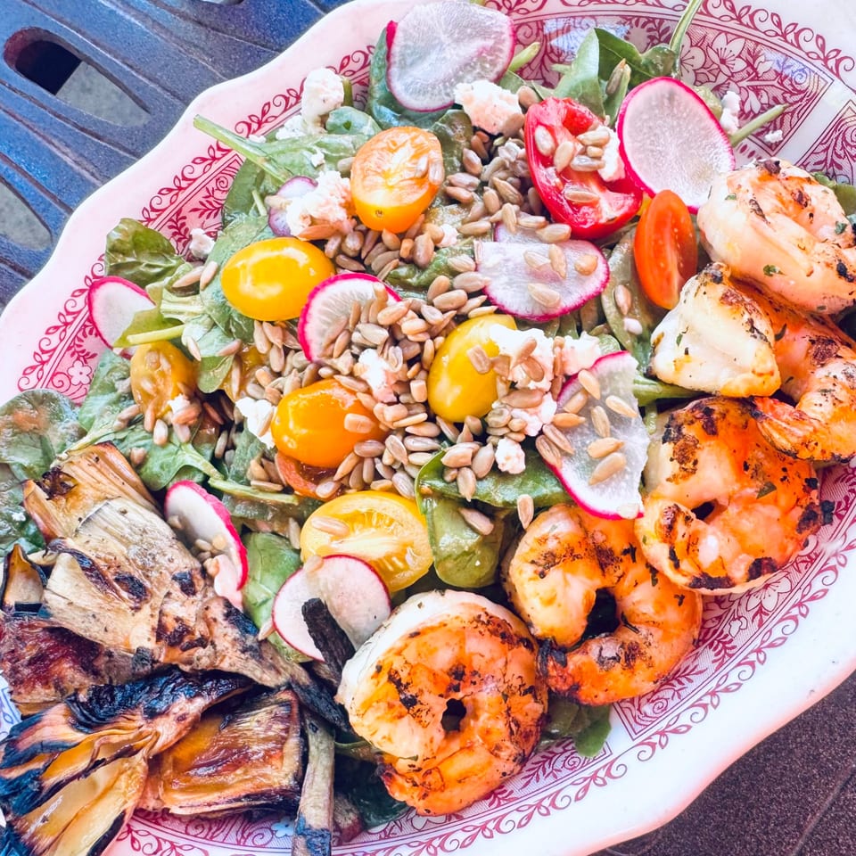 Grilled shrimp and vegetables, including radishes, tomatoes, and greens, on a decorative plate.