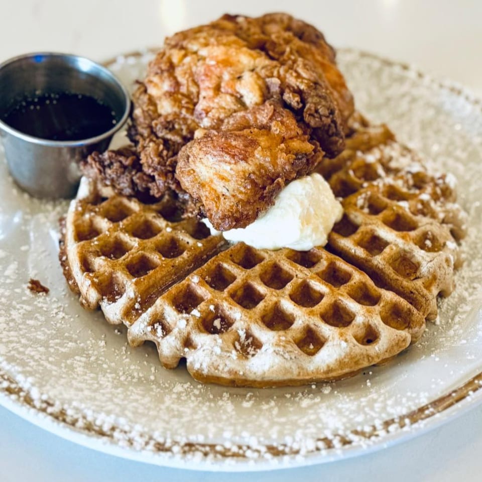 Chicken and waffles with syrup and whipped cream, dusted with powdered sugar on a white plate.