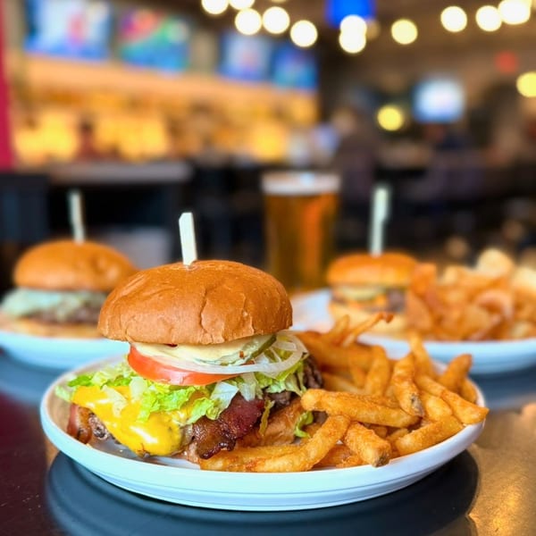 A Label Kitchen + Bar cheeseburger with bacon, lettuce, tomato, onion, and fries, with a beer in the background.