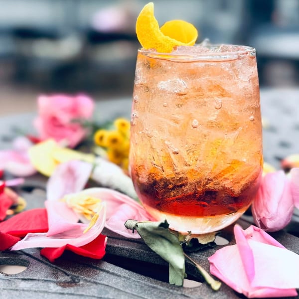 A Label Kitchen + Bar cocktail with a lemon twist and ice, surrounded by colorful flower petals.
